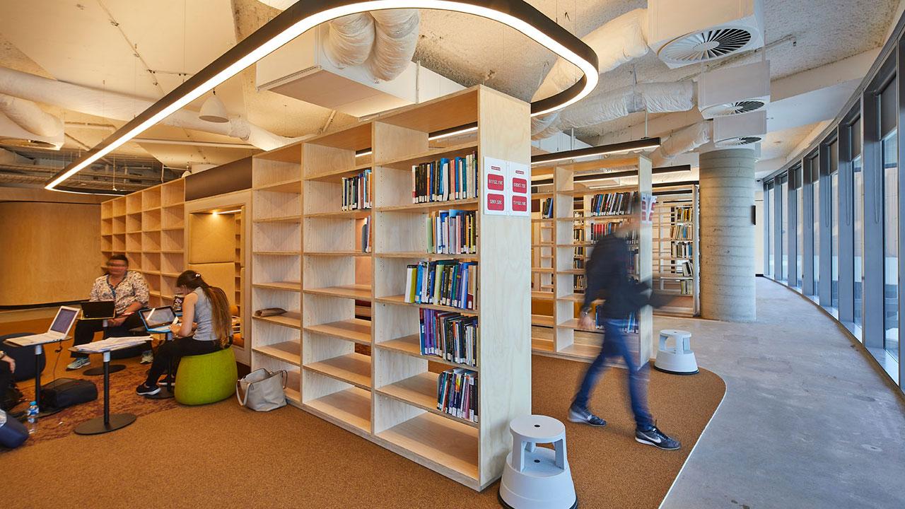 A university library. One student is walking from between the stacks in the middle ground. In the foreground students are seated with laptops